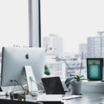 Un bureau propre et moderne équipé d’un Apple iMac, d’un ordinateur portable, d’une orchidée en pot et offrant une vue sur un paysage urbain à travers de grandes fenêtres.