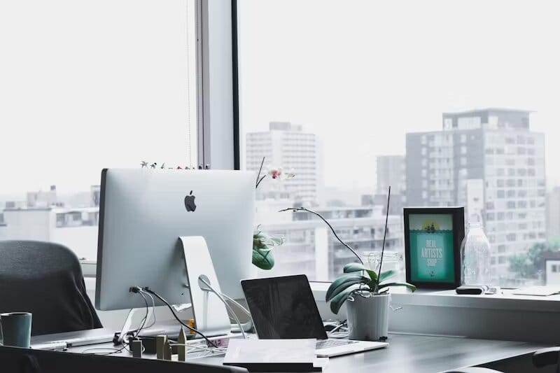 Un bureau propre et moderne équipé d’un Apple iMac, d’un ordinateur portable, d’une orchidée en pot et offrant une vue sur un paysage urbain à travers de grandes fenêtres.