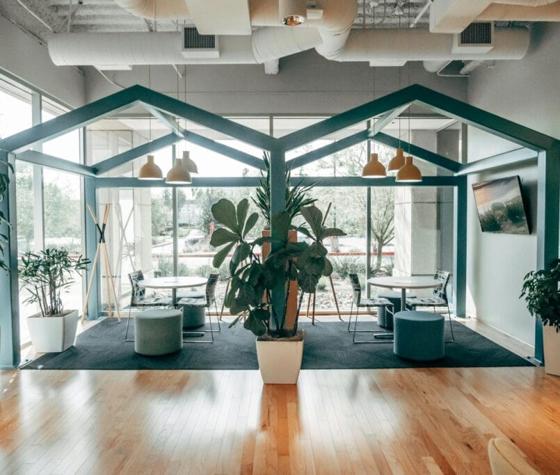 Un espace de coworking contemporain avec une zone de repos unique en forme de maison, des plantes vertes luxuriantes et de grandes fenêtres offrant une ambiance ouverte et aérée. Un éclairage suspendu et un sol en bois rehaussent le design moderne.