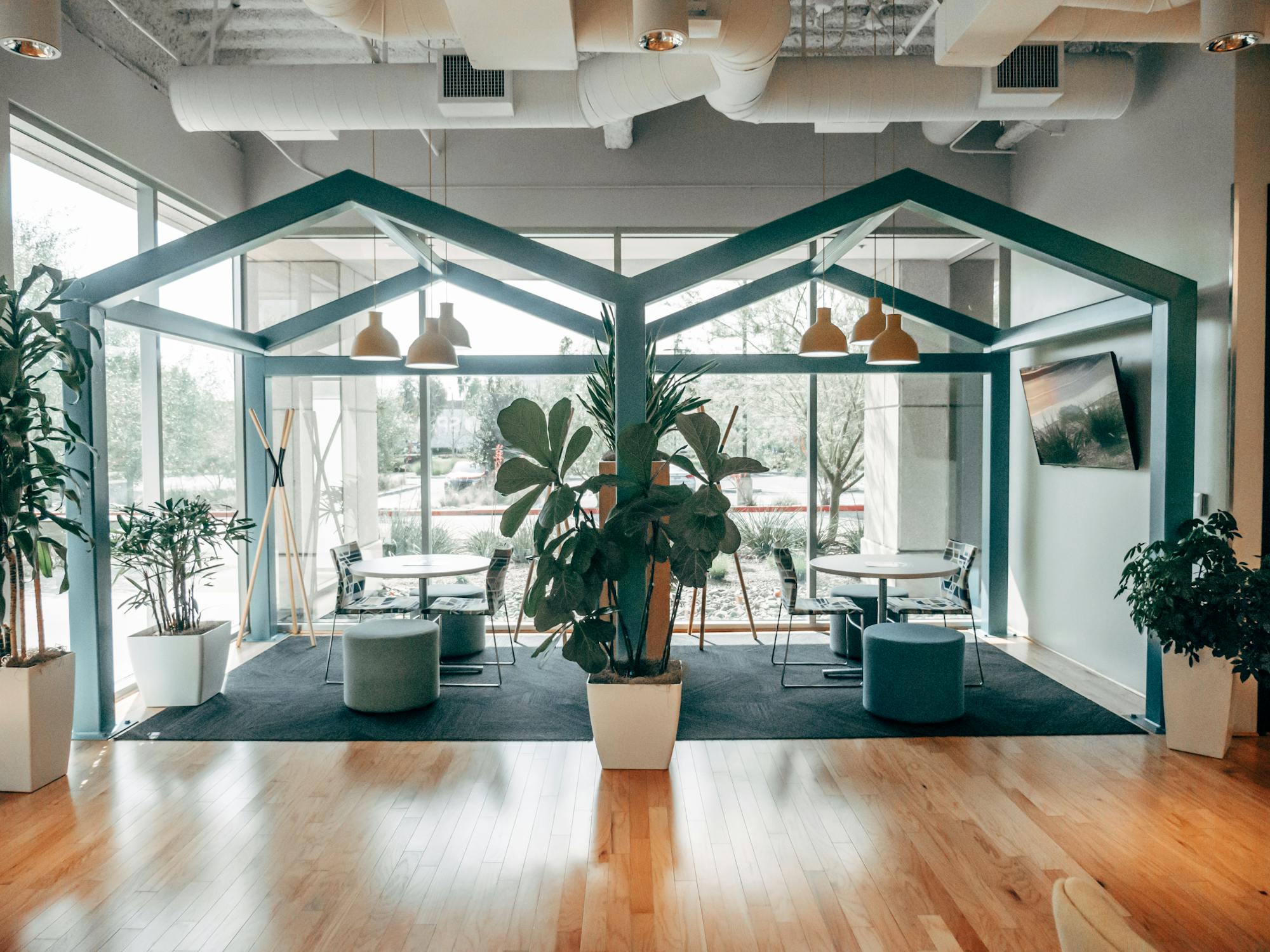 Un espace de coworking contemporain avec une zone de repos unique en forme de maison, des plantes vertes luxuriantes et de grandes fenêtres offrant une ambiance ouverte et aérée. Un éclairage suspendu et un sol en bois rehaussent le design moderne.
