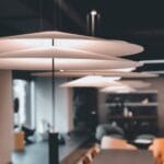 Vue en gros plan de luminaires suspendus minimalistes en forme de disque accrochés au plafond d’un bureau moderne. Les lampes circulaires blanches créent une esthétique épurée contrastant avec le plafond sombre, avec une vue floue sur une table de conférence et un espace de réunion en dessous.