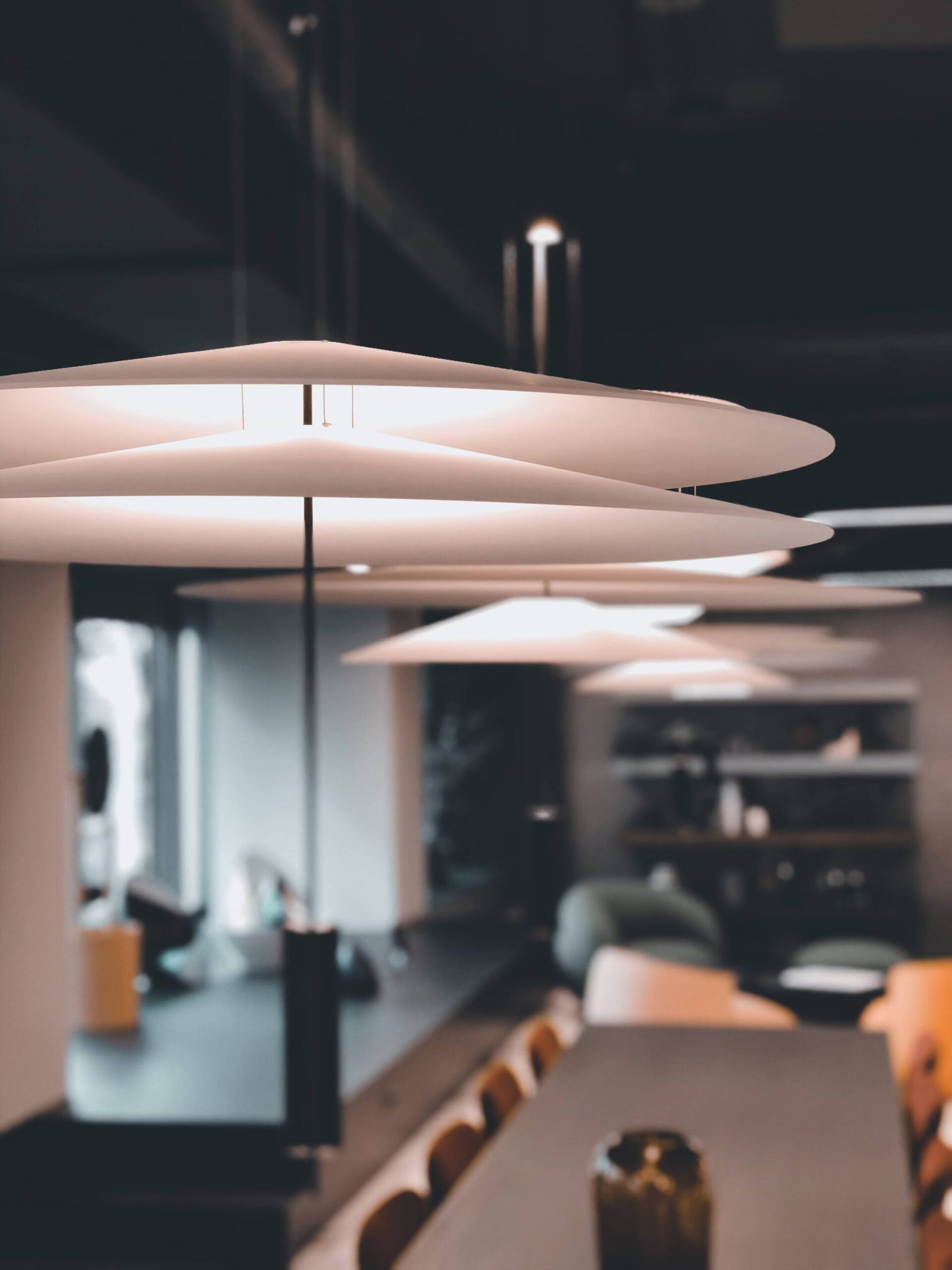 Vue en gros plan de luminaires suspendus minimalistes en forme de disque accrochés au plafond d’un bureau moderne. Les lampes circulaires blanches créent une esthétique épurée contrastant avec le plafond sombre, avec une vue floue sur une table de conférence et un espace de réunion en dessous.