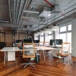 Espace de travail contemporain avec plafond en béton apparent, tuyauterie visible, lampes suspendues et parquet en bois en motif chevron, équipé d’ordinateurs et de chaises ergonomiques.