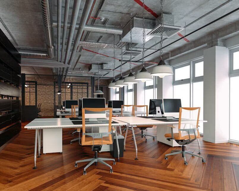 Espace de travail contemporain avec plafond en béton apparent, tuyauterie visible, lampes suspendues et parquet en bois en motif chevron, équipé d’ordinateurs et de chaises ergonomiques.