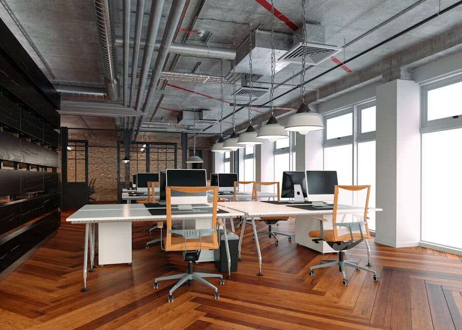 Espace de travail contemporain avec plafond en béton apparent, tuyauterie visible, lampes suspendues et parquet en bois en motif chevron, équipé d’ordinateurs et de chaises ergonomiques.