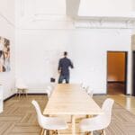 Salle lumineuse avec une grande table en bois clair, chaises blanches modernes, œuvres d'art au mur et une personne écrivant sur un tableau blanc.